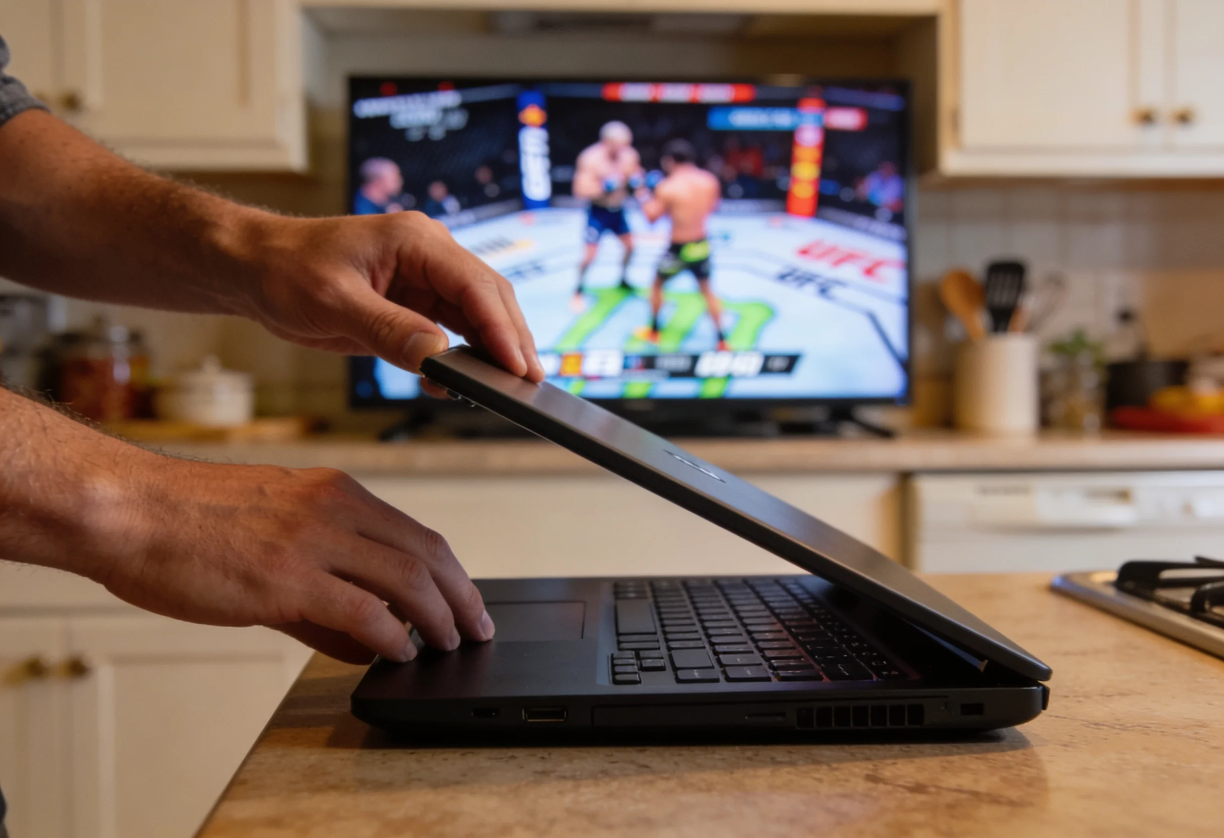 Person opening a laptop at a kitchen table with a UFC event visible on a TV in the background